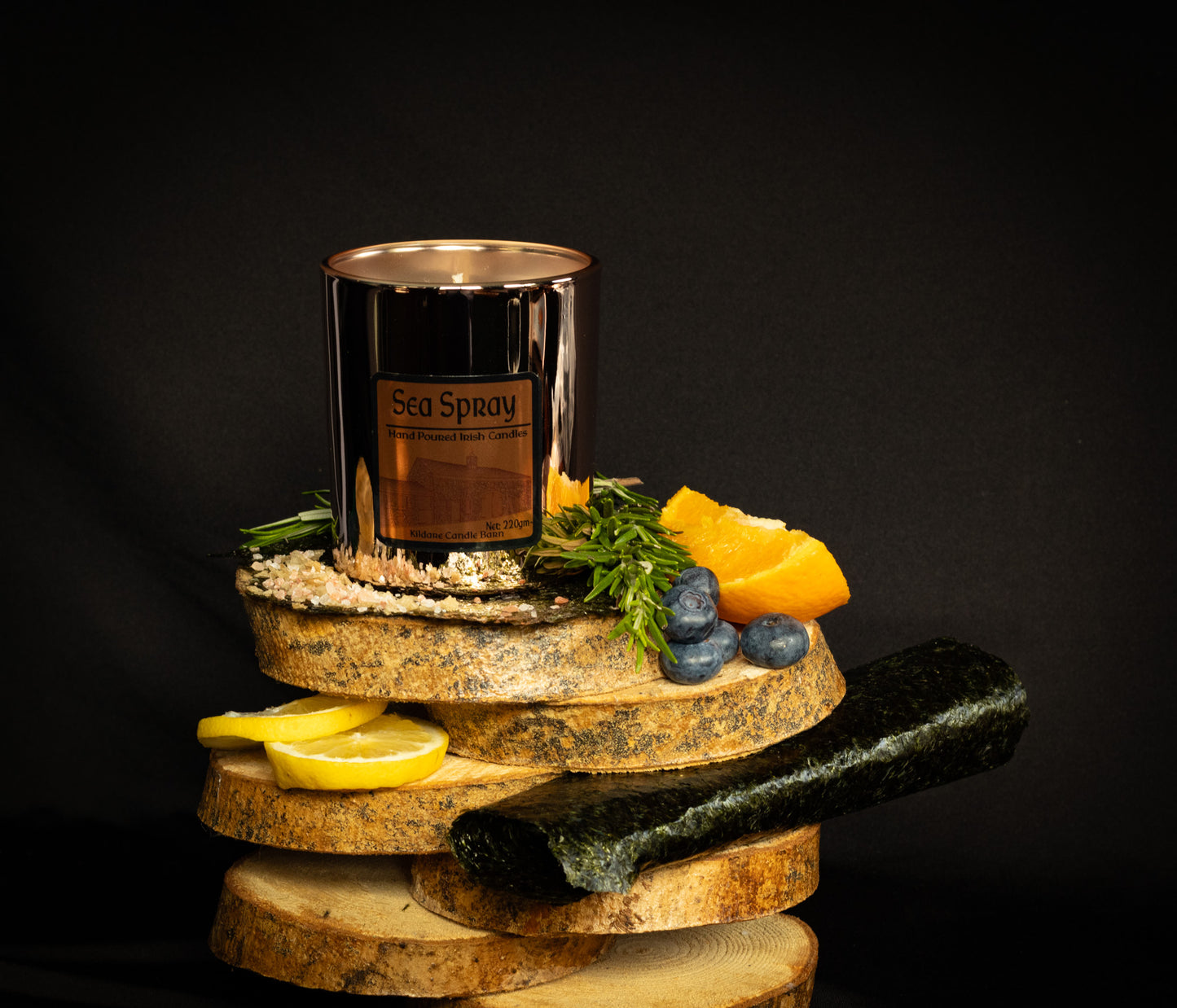 Irish Soy Wax Candle labeled 'Sea Spray'. Copper Jar Candle on a wooden display with lemons, stones, and herbs & seaweed against a dark background