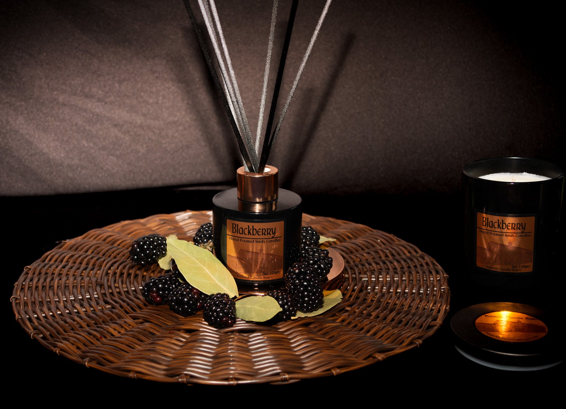 Blackberry-scented diffuser and candle on a woven tray with blackberries and bay leaves.