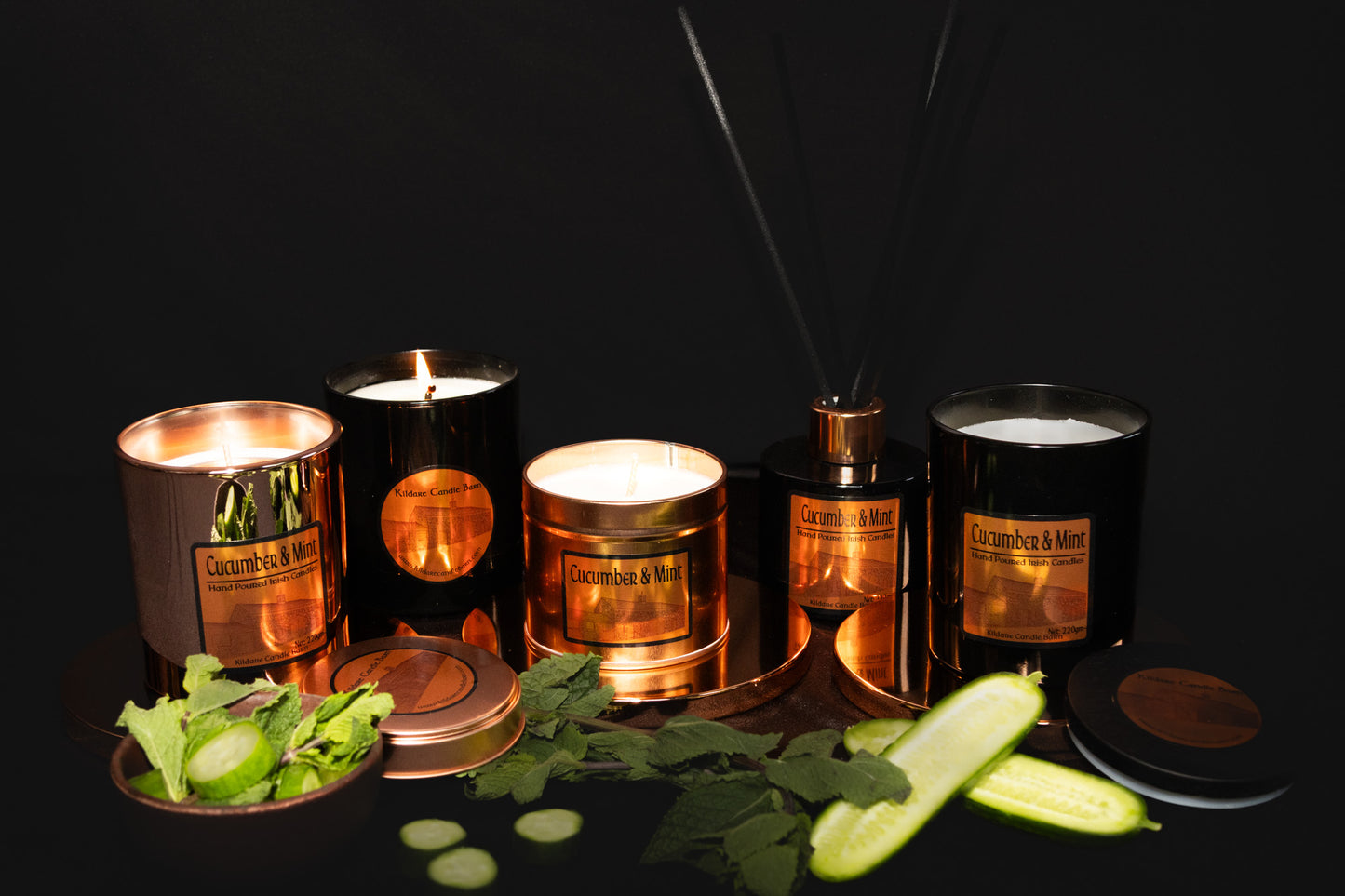 Collection of Irish Soy Wax Candles & Reed Diffusers labeled 'Cucumber & Mint' surrounded by sliced cucumbers and mint leaves on a dark background. Copper Tin