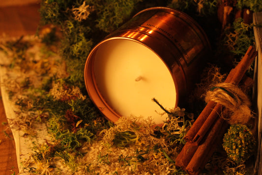 A soy wax candle with a copper lid on a bed of cedar and cinnamon sticks, surrounded by greenery and spices. Christmas Candle