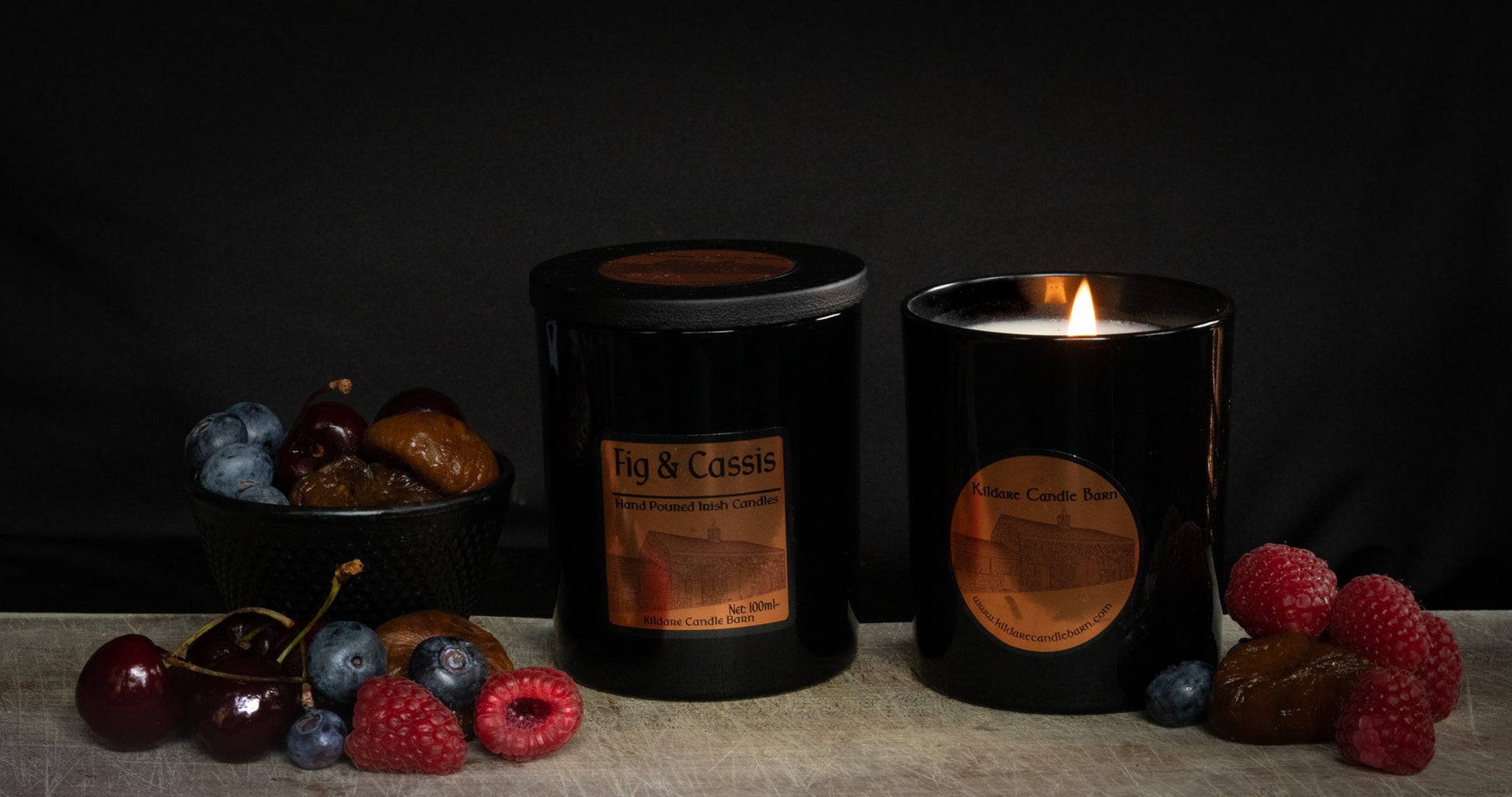 Fig & Cassis Irish Soy Wax candles in a black jar on a wooden surface with fruits of raspberry, cherry, fig and blueberry