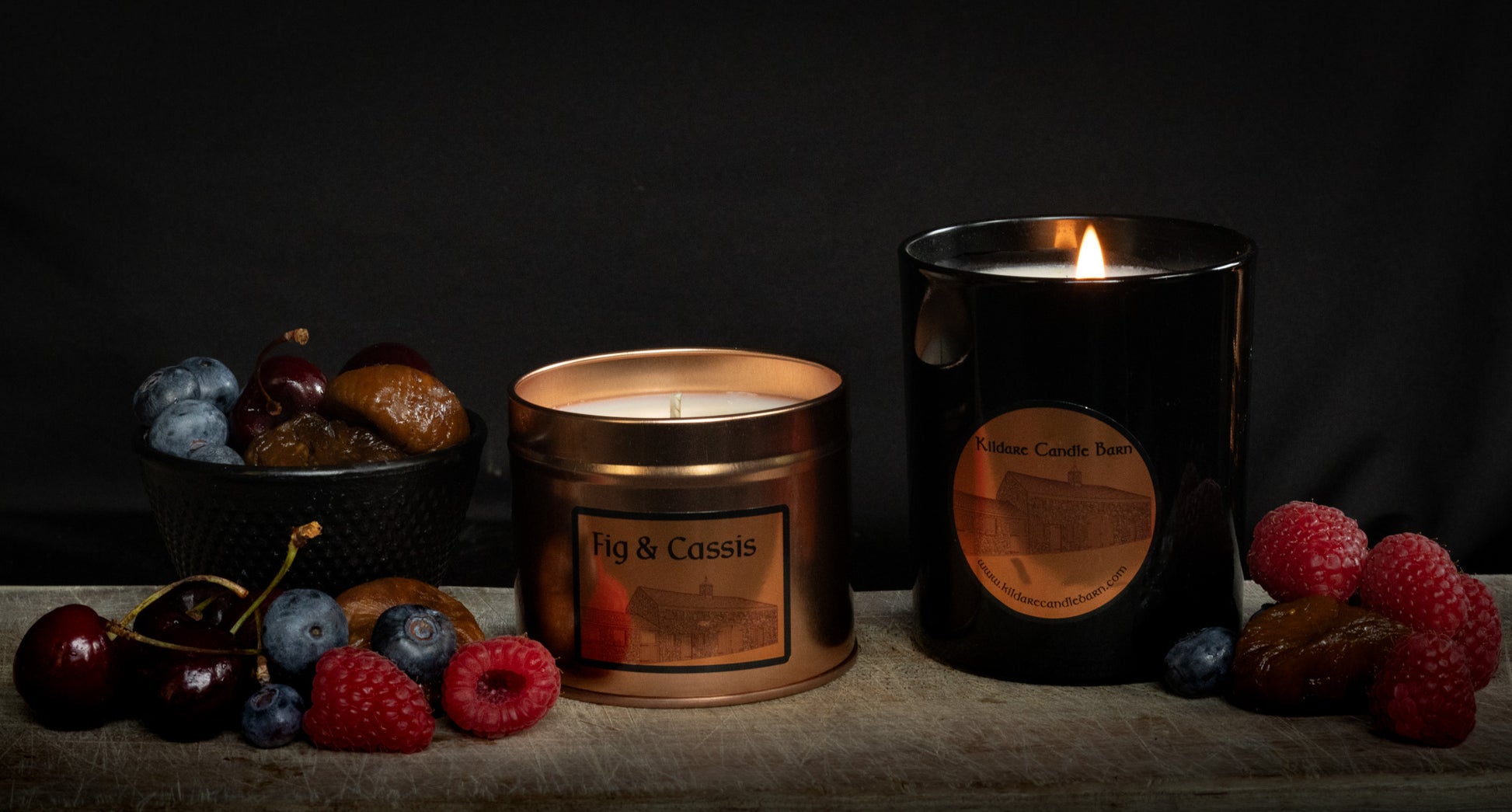Fig & Cassis Irish Soy Wax candles in a copper tin and a black jar on a wooden surface with fruits of raspberry, cherry, fig and blueberry