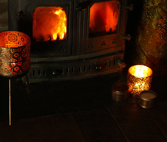 A lit soy wax candle with a speckled pattern on the lid, placed on a wooden surface in front of a fireplace.