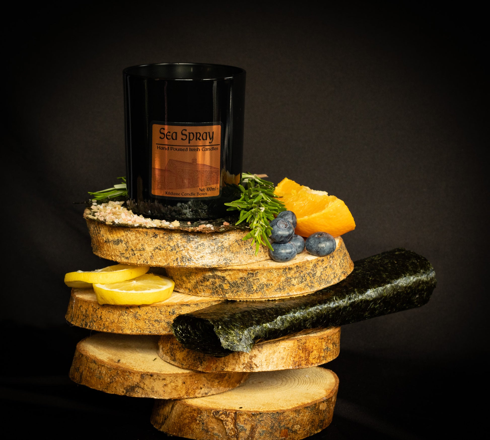 Irish Soy Wax Candle labeled 'Sea Spray'. Black Jar  Candle on a wooden display with lemons, stones, and herbs & seaweed against a dark background