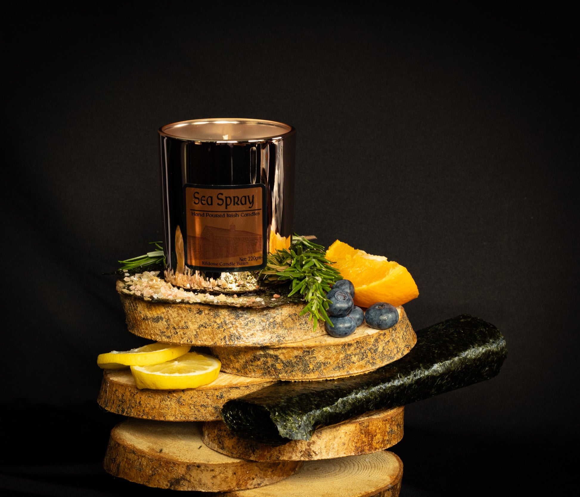 Irish Soy Wax Candle labeled 'Sea Spray'. Copper Jar  Candle on a wooden display with lemons, stones, and herbs & seaweed against a dark background