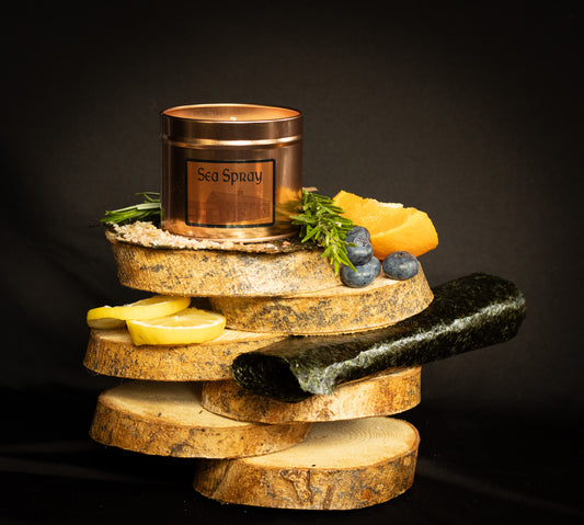 Irish Soy Wax Candle labeled 'Sea Spray'.Tin Candle on a wooden display with lemons, stones, and herbs & seaweed against a dark background