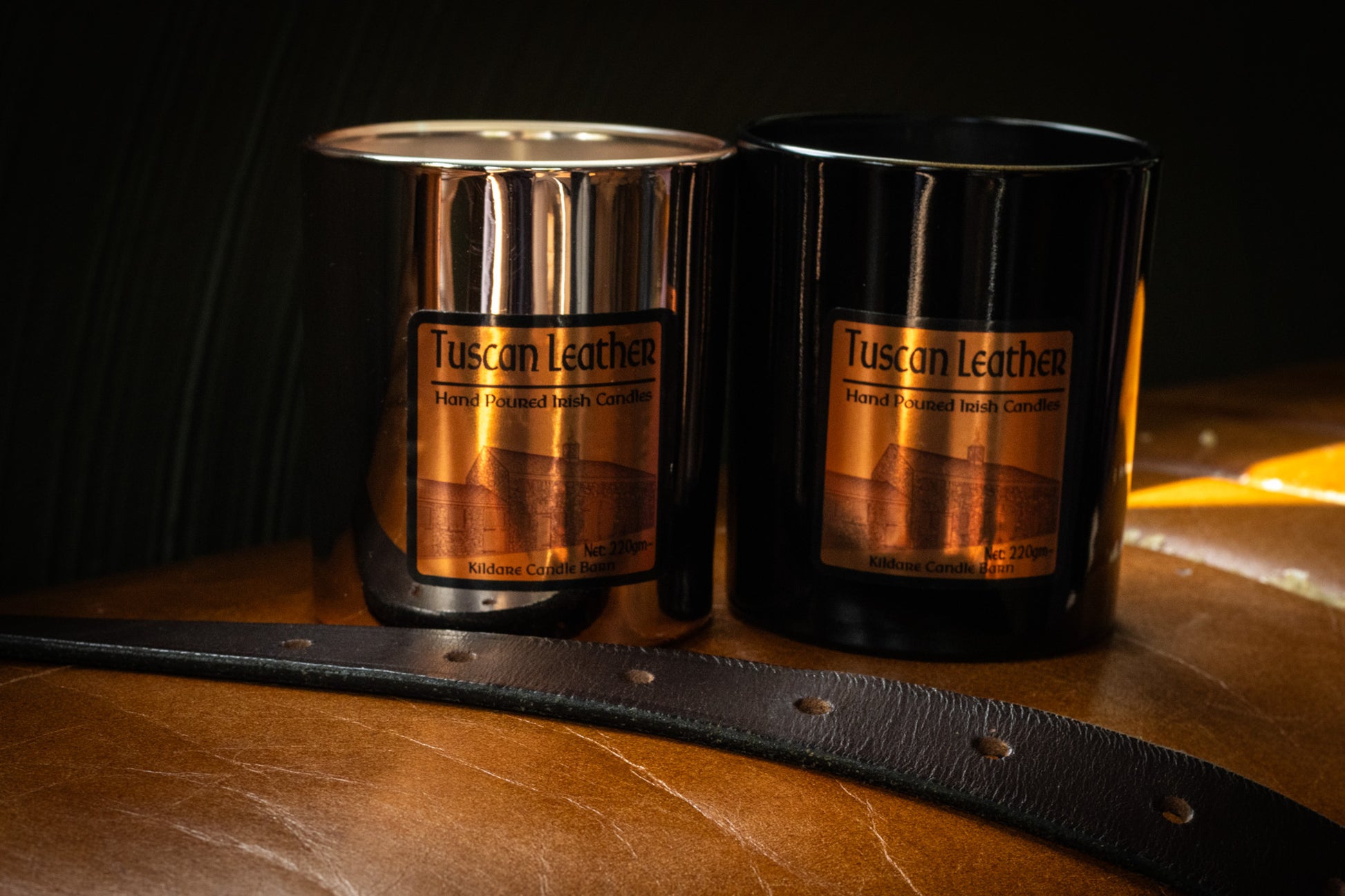 Two Tuscan Leather soy wax  candles on a wooden surface with a leather strap.