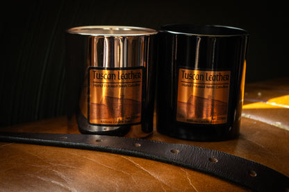 Two Tuscan Leather soy wax  candles on a wooden surface with a leather strap.