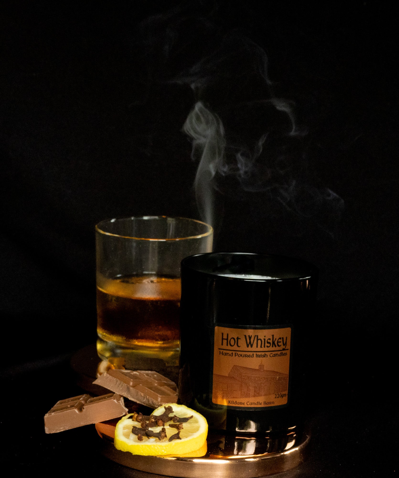 Irish Soy Wax Candle in a Black Jar with a Hot whiskey glass with a lemon slice and smoking ambiance on a dark background
