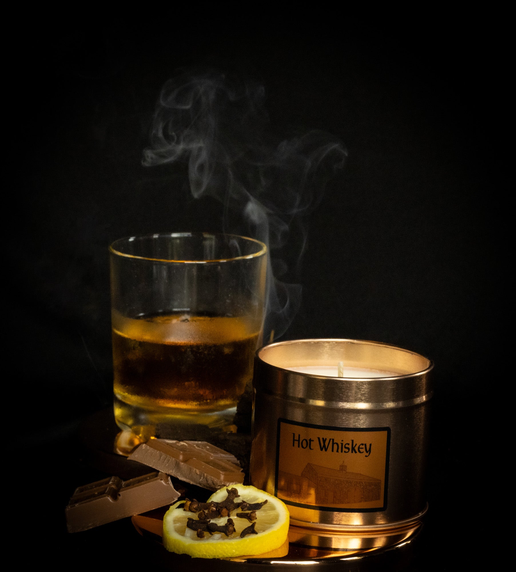 Irish Soy Wax Hot whiskey candle in a copper tin with a glass of whiskey and chocolate and clove studded lemon on a dark background